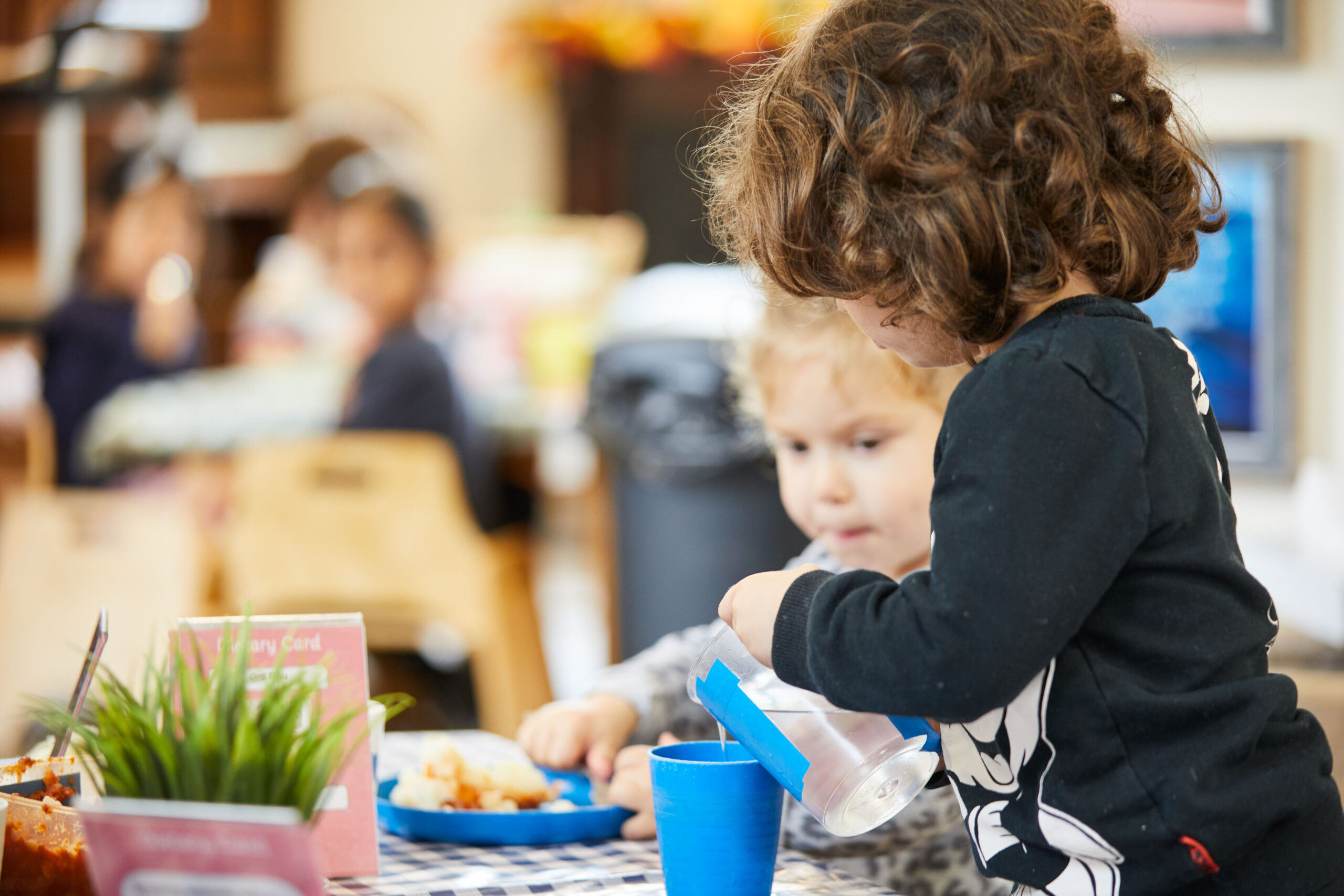 2 nursery children at the dining table with one pouring some water into a cup