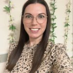 Young woman with long brown hair and round glasses smiles at camera while taking a selfie; she's wearing a beige floral blouse against a wall adorned with vertical green-and-white flower garlands.