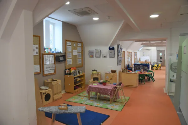 Small wooden children's table arranged with toy dishes, surrounded by chairs; sits on a rug amid shelving and activity stations in a bright, orange-floored preschool classroom corridor.