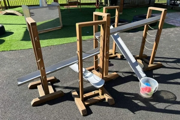 Wooden support frames holding angled metal troughs guide colorful balls down into a plastic bucket on asphalt, in an outdoor playground area with turf and a fence.