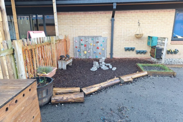 Small wooden climbing wall displays colorful holds mounted against a brick building; a mulch-covered play area contains stacked cinder blocks, curved log borders, planters, and a wooden fence.