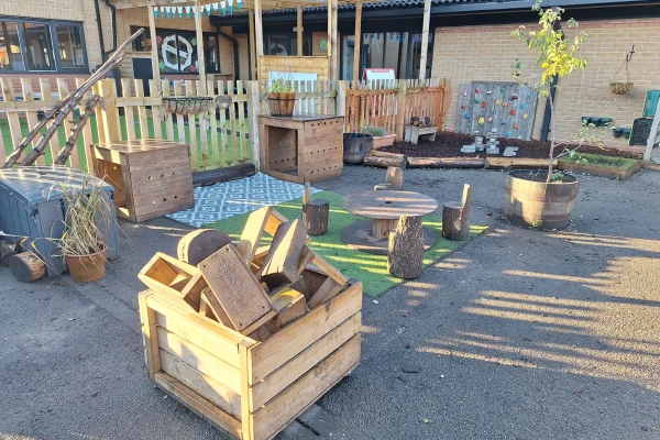 Wooden crate filled with wooden blocks sits among stump seats and a spool table in an outdoor preschool play area with planters, a climbing wall, low fence, and building backdrop.