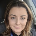 Woman's face, looking directly at the camera with a slight smile, seated in a car interior with headrest and seatbelt visible; lanyard reads 