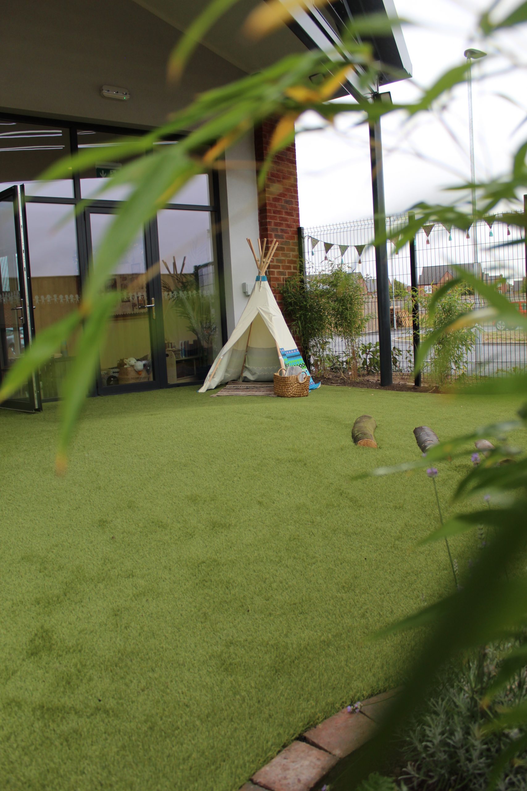 Small canvas teepee sits open on green artificial turf beside a wicker basket near glass patio doors and a brick column, framed by leafy plants and a fenced outdoor courtyard.