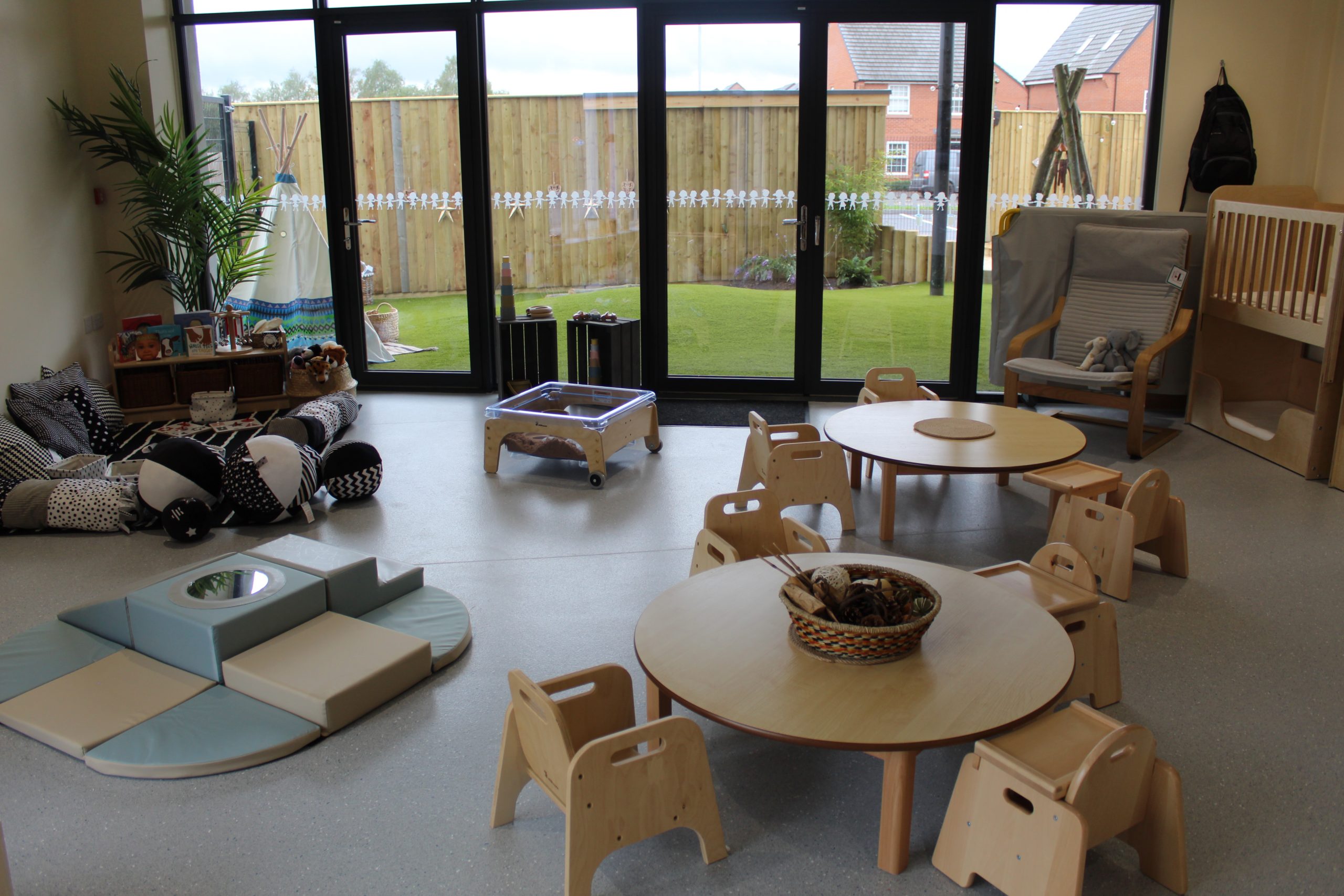 Round wooden tables surrounded by small chairs, basket of natural materials and soft play cushions; toys scattered in a bright classroom with glass doors opening to a fenced grassy yard.