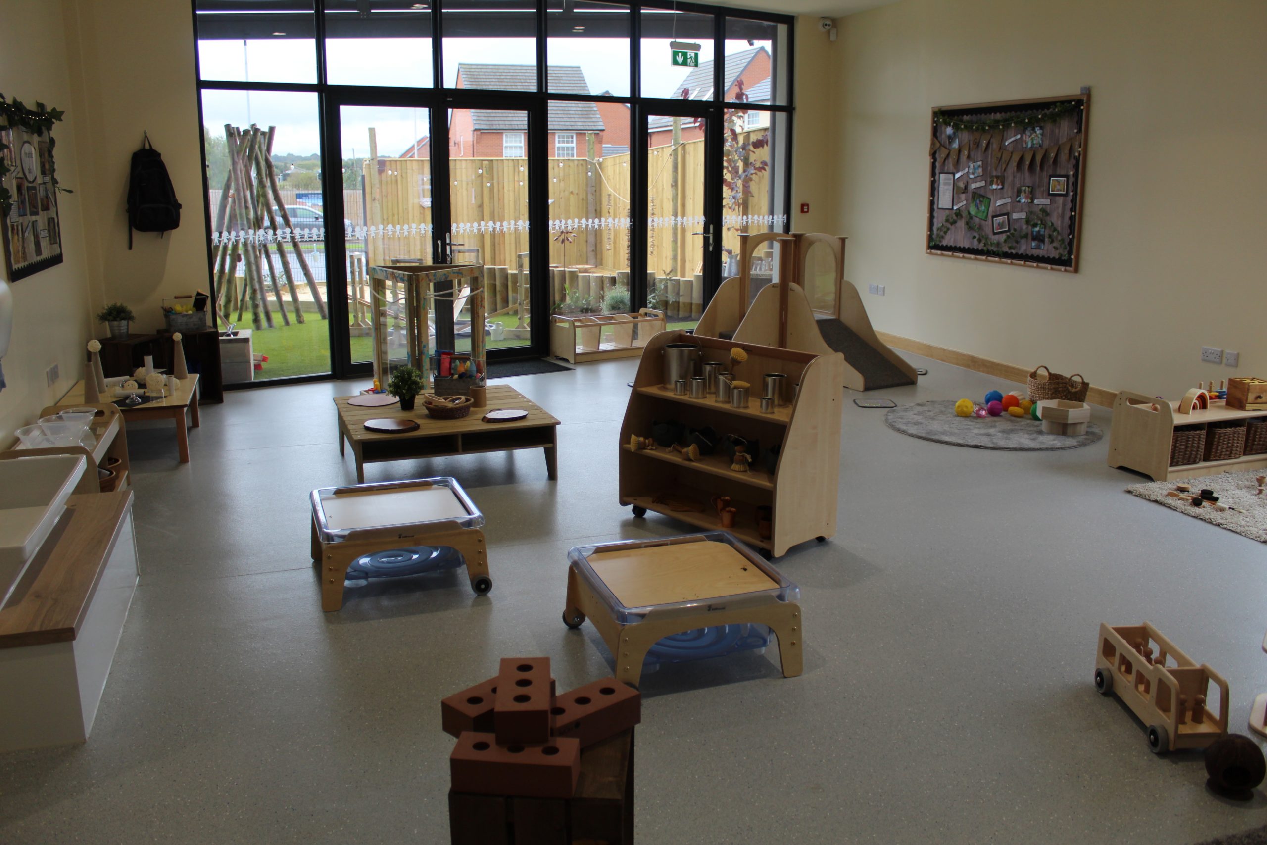 Small wooden play furniture and toys are arranged across a bright preschool room, clustered into play stations near large glass doors that open onto a fenced outdoor play area.