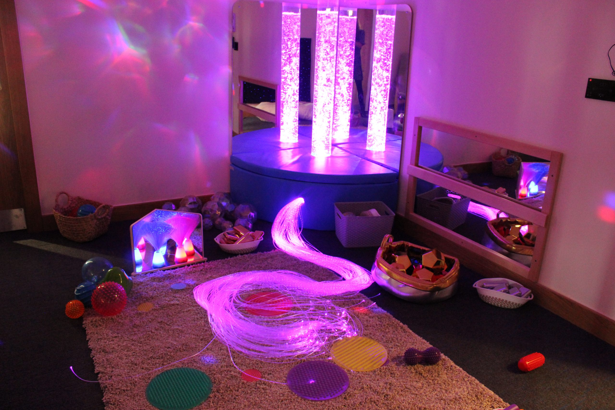 Fiber-optic light strands glow purple, coiled on a shag rug; in a sensory room corner with mirrored panels, illuminated bubble tubes, round cushions, baskets, and scattered tactile toys.