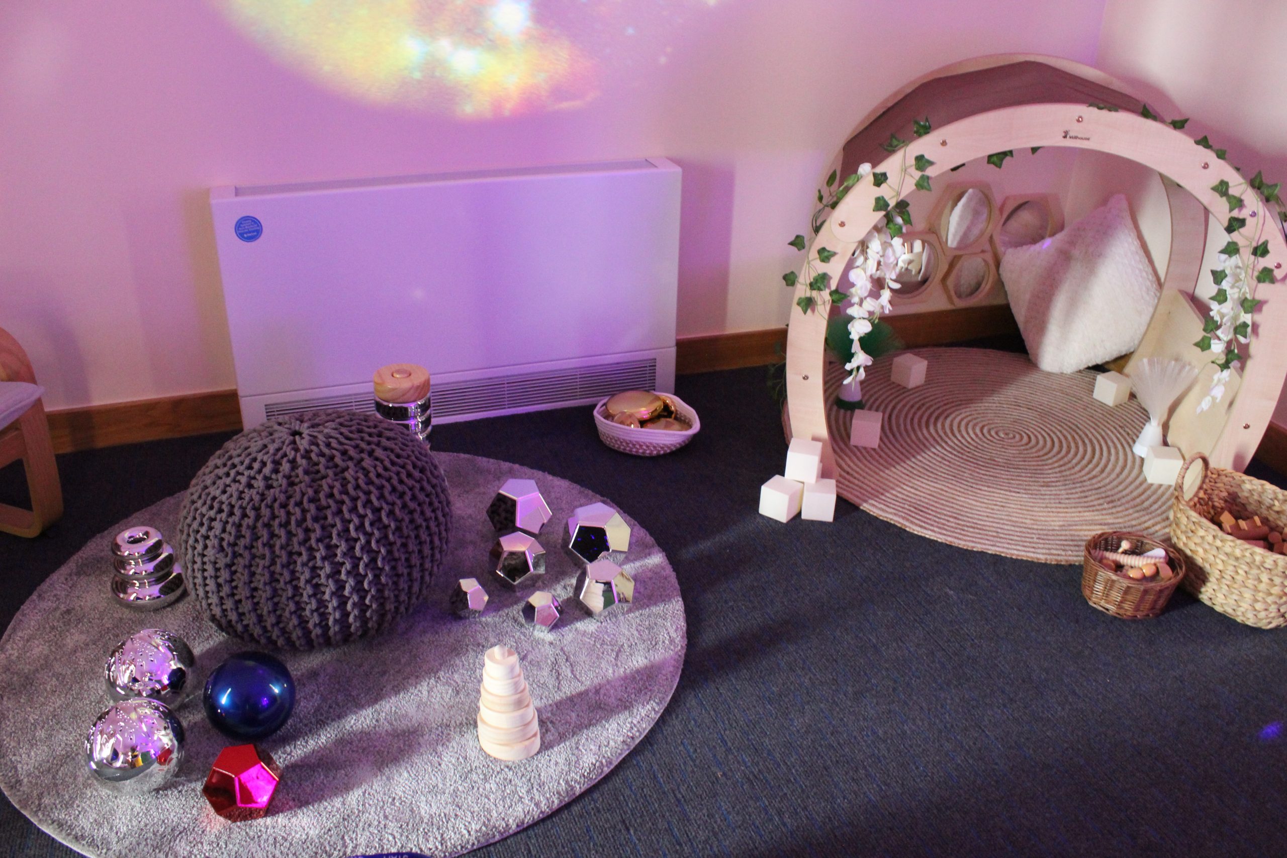Knitted gray pouf rests on a round rug, surrounded by shiny geometric toys; a wooden arch draped with faux ivy and cushions frames cozy play corner against a pink-lit wall.