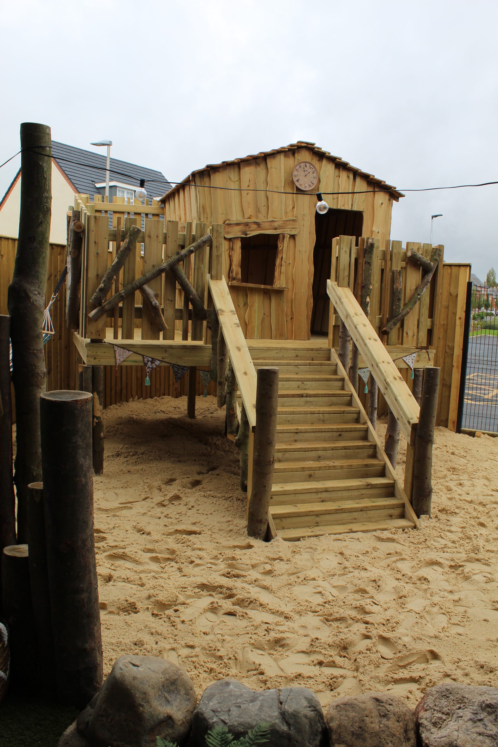 Wooden playhouse elevated on stilts with log-rail stairs leading up; small window and clock above the entrance. Surrounded by deep sand, a wooden fence and nearby houses under a cloudy sky.