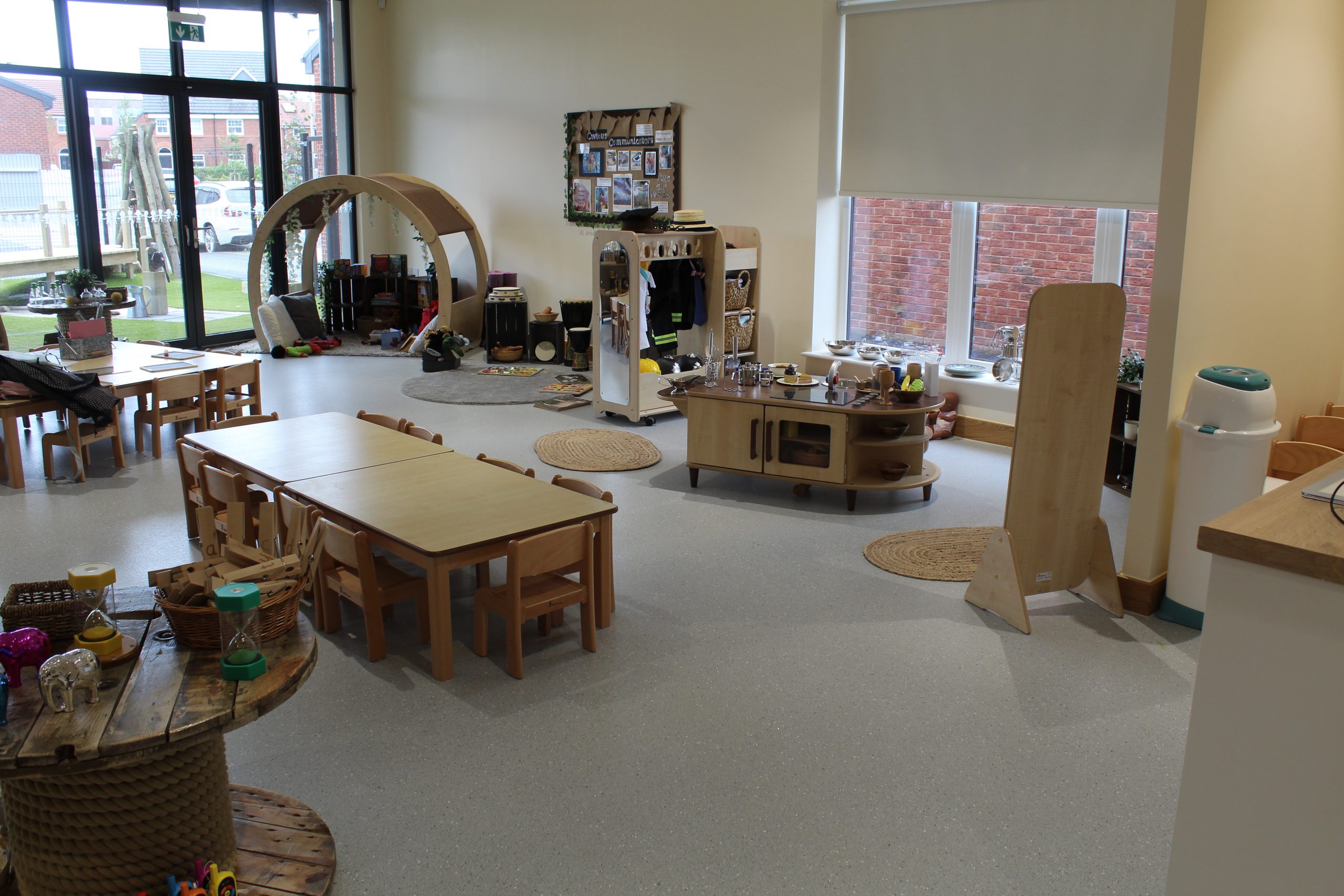 Small wooden tables and chairs sit arranged for children in a tidy preschool; a play kitchen, reading nook with circular arch, rugs and toys fill a bright room with windows.