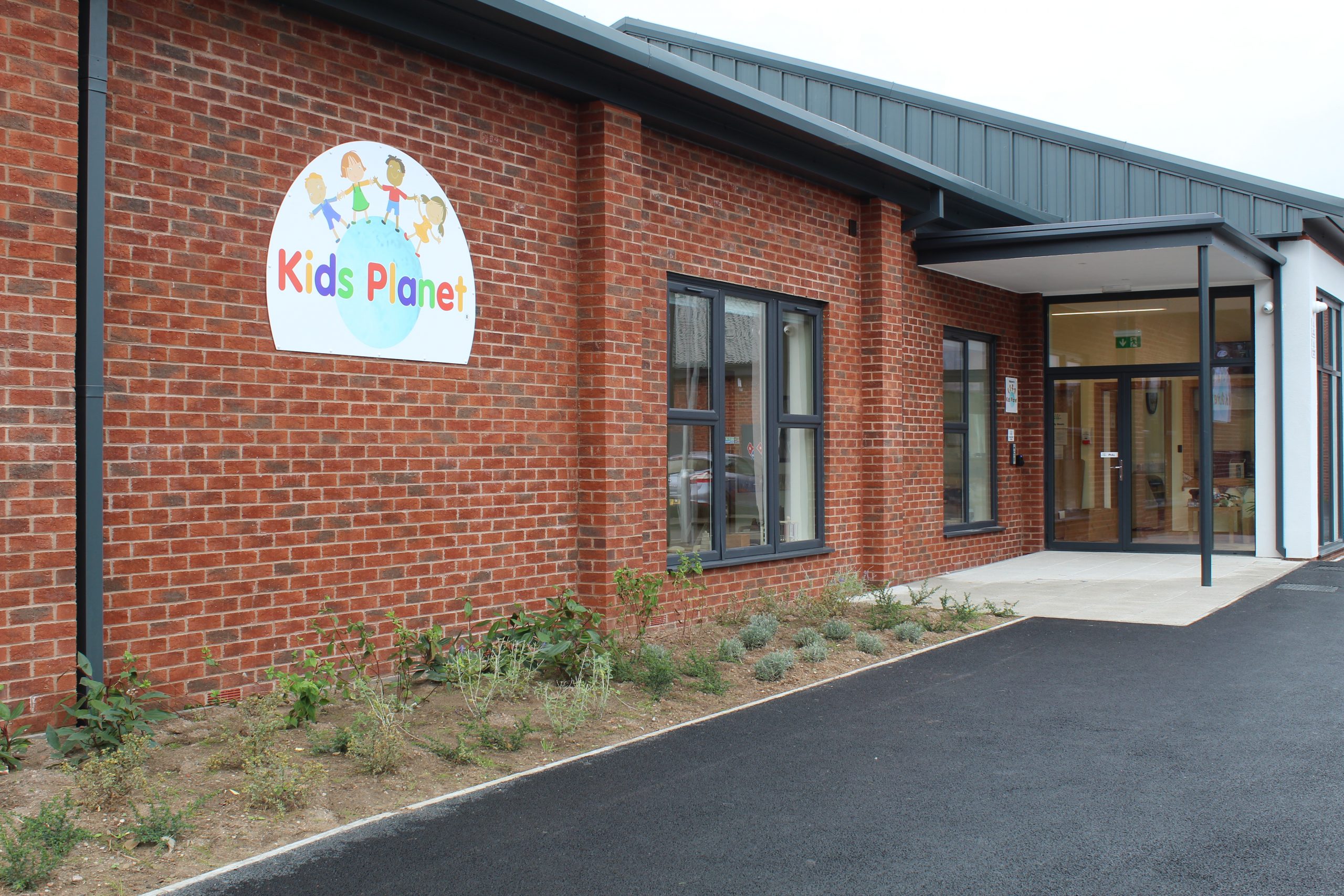 Brick childcare building displays colorful sign reading "Kids Planet" with cartoon children above a globe; glass entrance doors under a canopy, small planted border and asphalt drive in front.