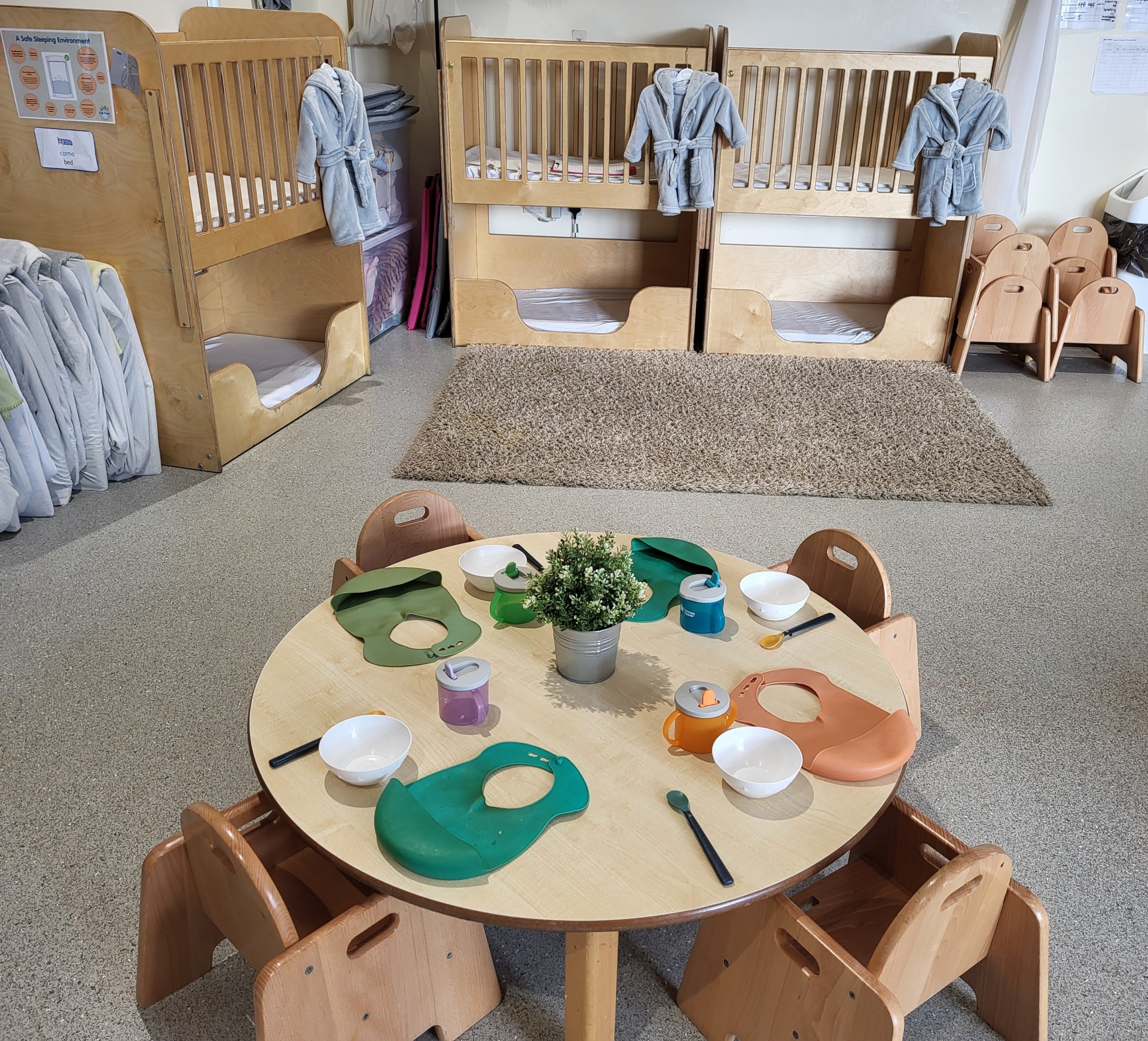 Round children's table set with silicone bibs, bowls, sippy cups, spoons and a small potted plant, arranged for a meal in a daycare room with wooden cribs, hanging robes and a rug.