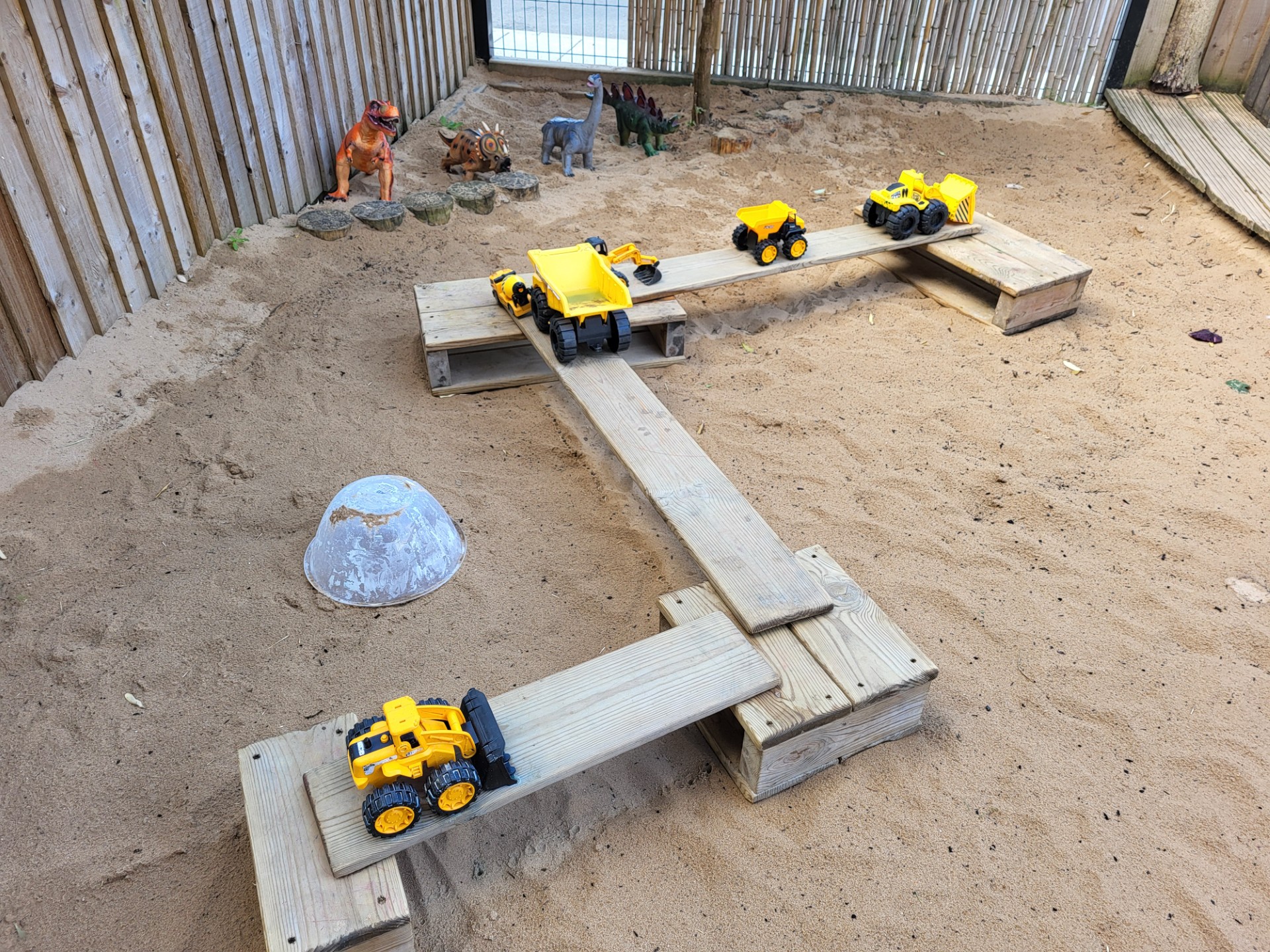 Yellow toy construction vehicles sit on wooden plank ramps and platforms inside a sandy play area, flanked by plastic dinosaur figures along a wooden fence and an overturned white bowl.