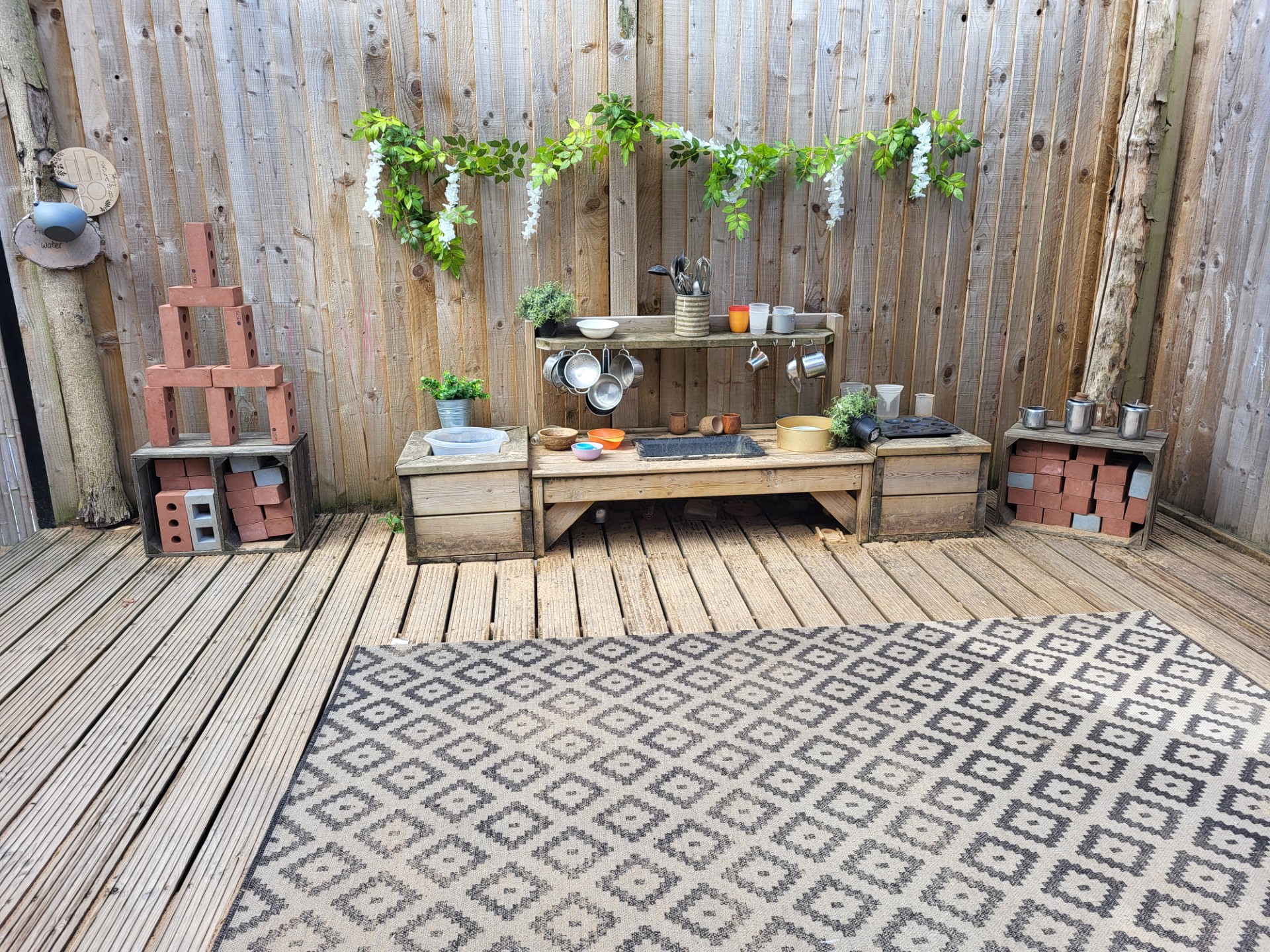 Low wooden mud-kitchen bench holds hanging metal pots, cups, bowls, potted plants, and stacked bricks. Against a vertical wooden fence on a deck with a patterned outdoor rug.