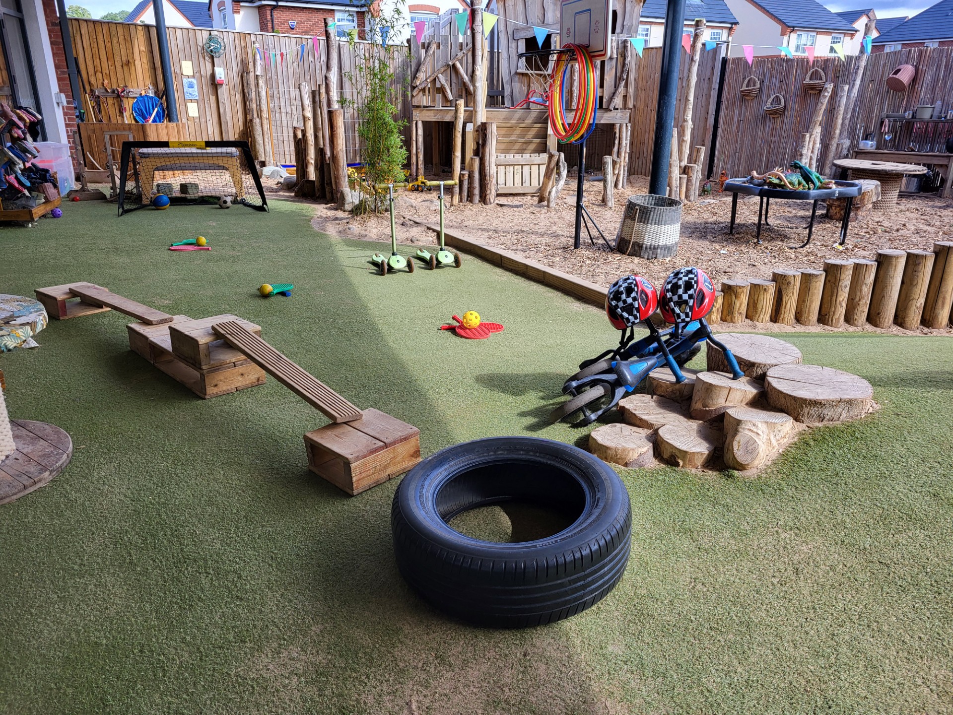 A black tire sits on artificial turf, surrounded by wooden balance beams, stepping logs and scooters; two children's helmets rest on a fallen tricycle in a fenced outdoor playground.