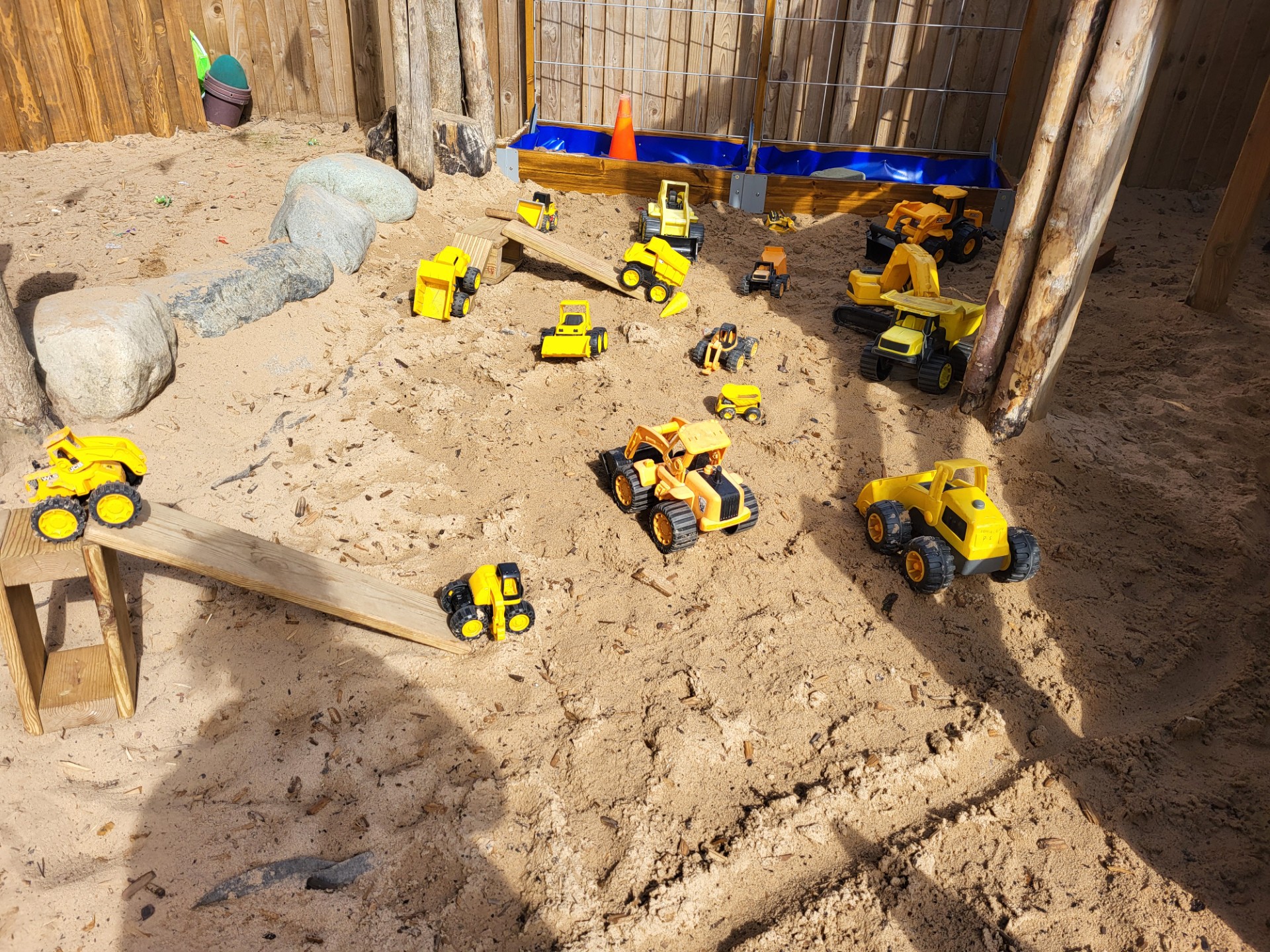 Toy yellow construction vehicles scattered across a sandy sandbox, some driving up wooden ramps and parked near wooden posts and rocks within a fenced play area.