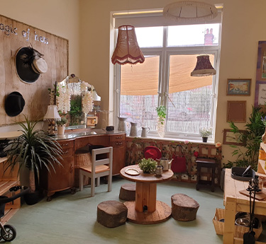 Small round wooden spool table surrounded by stump-like cushions, holding a plant; vintage wooden cabinets, hats and lamps arranged beneath a sunlit window in a cozy classroom-like corner.