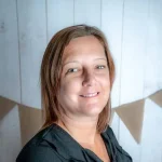 Middle-aged woman with shoulder-length light brown hair, smiling gently while slightly turned to camera; wearing a dark blouse against a light wooden plank wall decorated with triangular burlap bunting.
