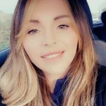 Smiling woman looks at camera, seated in a car; long light-brown highlighted hair frames her face, wearing a dark top, with daylight and car headrests visible through the windows.