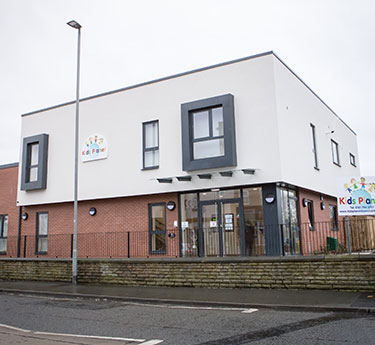 Two-story childcare building stands beside a wet street, with white upper walls, red-brick lower facade, boxy window bays, and a glass entrance under an overcast sky.