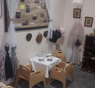 Child-sized table set for tea, four small chairs arranged around it; toy teapot and cups on a cloth. In a playroom corner with hanging hats, dresses, bulletin board and curtains.