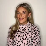 Young woman smiling at camera against a plain light wall, wearing a pink high-neck blouse with black abstract spots; long blonde hair draped over one shoulder.
