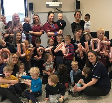Rose-gold letter balloons spell "CONGRATULATIONS" as caregivers and toddlers pose together on a carpeted daycare classroom floor, adults standing or kneeling while holding children.