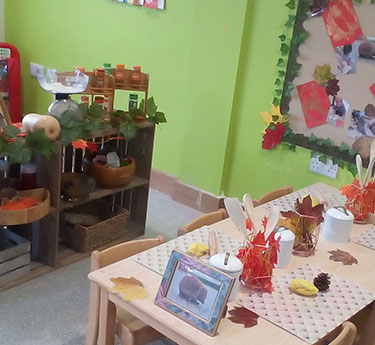 Small wooden table displays autumn place settings—paper leaves, pinecones, crafted lanterns, cups and a framed photo—arranged for children in a bright green preschool room with shelves and a bulletin board.