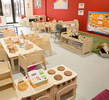 Low wooden play kitchen holds a toy cash register and play food; nearby small tables and chairs set for activities; bright preschool classroom with colorful walls, shelving, and soft rug.