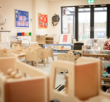 Small wooden chairs and tables sit arranged around activity stations, with toys, storage units, colorful bulletin boards, and glass exit doors in a bright, organized preschool classroom.