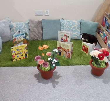 Children's books stand upright on a small patch of artificial grass; cushions and a stuffed dog sit behind them, two potted flowers in front, and a low bookshelf to the right.