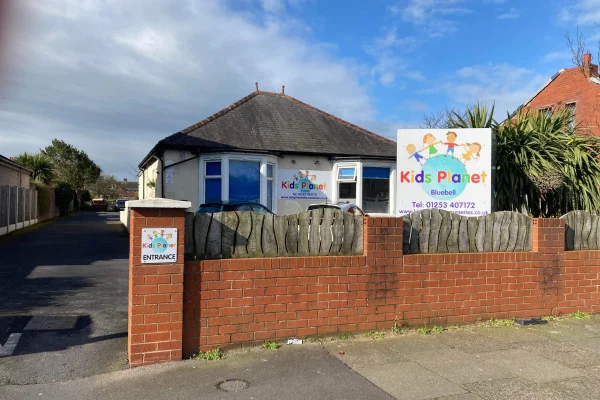 Colorful "Kids Planet" nursery sign stands on a brick fence as a single-story bungalow sits behind; driveway to the left, palm-like shrubs to the right, partly cloudy blue sky overhead.
