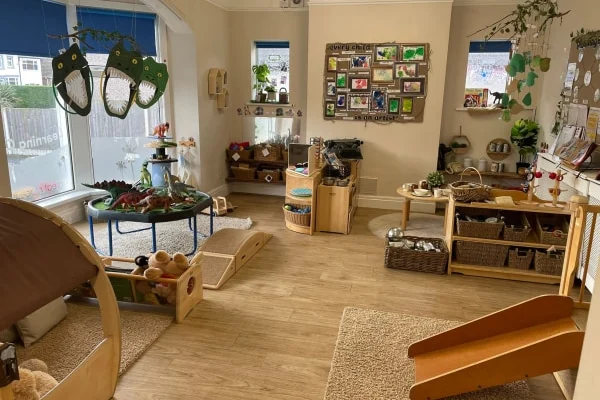 A preschool playroom: wooden play structures and low shelves arranged for exploration, scattered toys and baskets, soft rugs and hanging decorations, large windows letting in natural light.