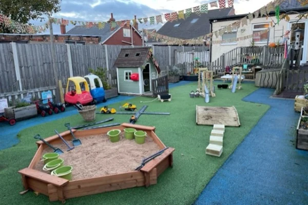 Wooden sandbox holds sand, shovels, and green buckets; toy cars and small playhouse sit nearby on artificial turf, bunting strung overhead in a fenced outdoor play area.