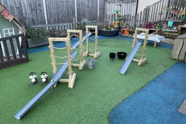 Three wooden ramp frames with gray plastic chutes form sloping channels, arranged for rolling or pouring; set on green artificial turf in a fenced outdoor play area with toys.