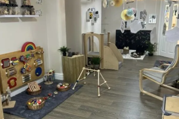 Wooden activity panel mounted low on left wall, with baskets of toys on a rug; small wooden climber and play structure sit on laminate floor amid chairs and hanging decorations.