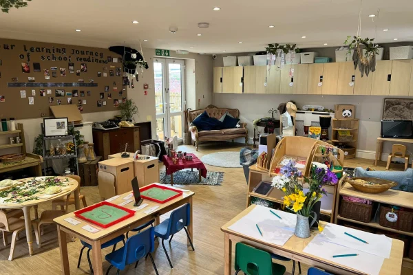 Early-years classroom — tables and chairs arranged with art materials, play areas and a sofa; children’s learning displays on the wall. Text visible: "our learning journey of school readiness" and "EXIT".