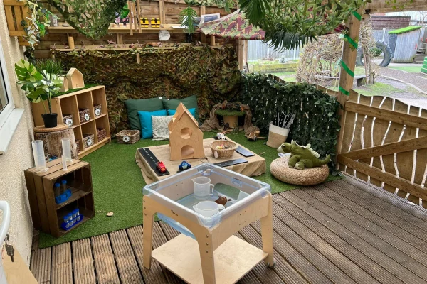 Low wooden sensory table holds water, cups and a leaf; sheltered outdoor children's play corner features artificial grass, cushions, toy house, wooden crates, plants and decking.
