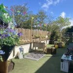 Wooden bench and cushioned chair sit on artificial turf, surrounded by potted and hanging flowering plants and decorative crates, under a sunny blue sky with trees behind a wooden fence.