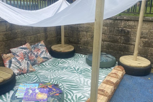A cozy outdoor reading nook: floor cushions and patterned pillows arranged on a leafy rug, children's books stacked on a low table, under a white canopy in a walled corner.