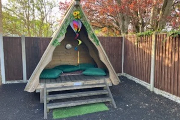 A wooden A-frame shelter sits on a raised platform, furnished with green cushions and a step; a colorful hanging spiral decoration dangles inside; fenced yard and autumn trees surround it.