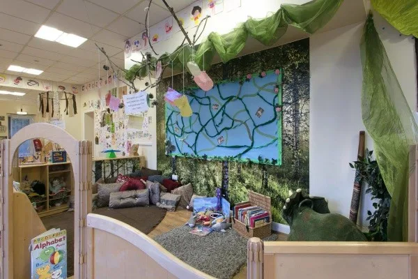 Cozy reading nook shows floor cushions and children's books arranged beneath hanging paper lanterns and green fabric vines in a nature-themed classroom corner. Text: "Alphabet"