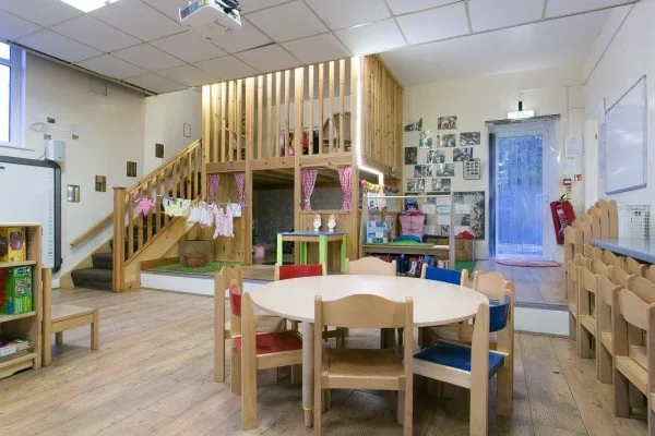 Wooden loft play structure creates a raised play area; a small clothesline with toy garments hangs from the railing. Low round table and colorful chairs fill a bright preschool classroom.