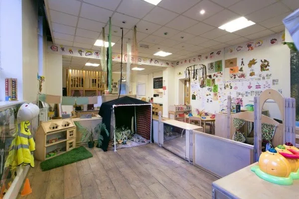 A black play tent sits open, inviting children; surrounded by low wooden shelves, toys, craft stations and colorful artwork along walls in a bright, organized daycare classroom.