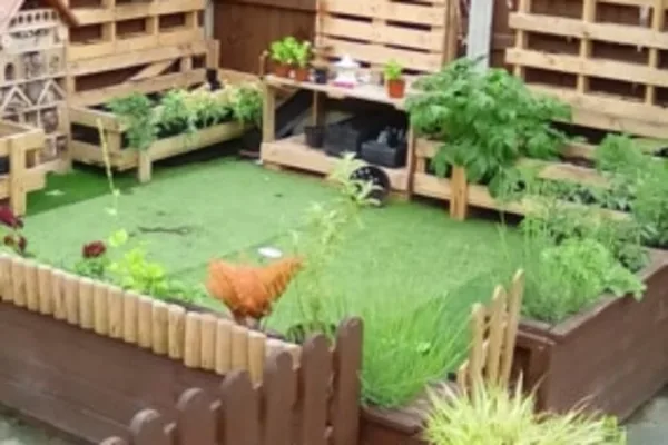 Brown hen walks on artificial grass inside raised wooden planters, surrounded by small potted herbs, pallet fencing and a low picket border in a compact backyard garden.