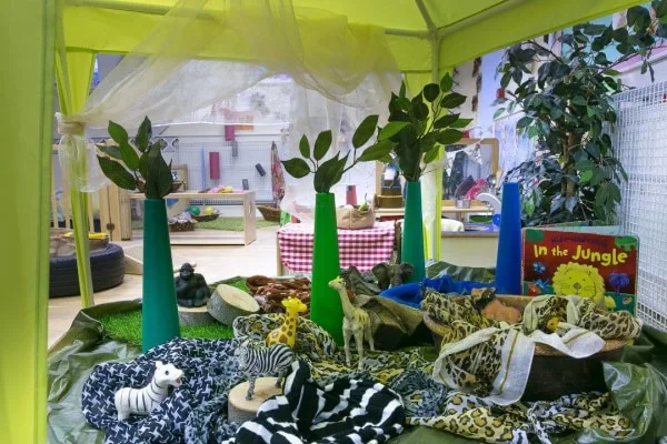 Toy jungle animals (zebras, giraffes, snakes) are arranged among fabric "grass" and cone "trees" under a yellow canopy; indoor playroom setting with plants and a book titled "In the Jungle".