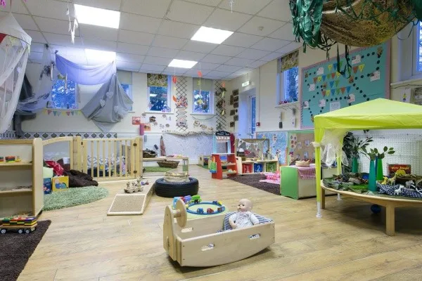 Wooden toy boat holds a baby doll, stationary on the floor; preschool playroom surrounds it with rugs, cushions, low shelving, activity stations, soft tents, windows, and scattered toys.