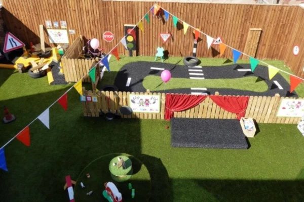 Miniature fenced play-road features toy road markings, tires, and a pink balloon; bunting lines cross above on artificial turf in a backyard play area. Text: "STOP"
