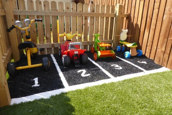 Four children's ride-on toys sit in numbered parking stalls on black mulch beside a wooden picket fence, facing a patch of artificial grass in a sunny fenced yard. Text: "1 2 3"