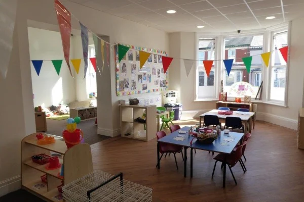 Colorful triangular bunting flags hang across a bright preschool classroom, overlooking small tables and chairs with craft materials, toy shelves, a bulletin board, and large sunlit bay windows.
